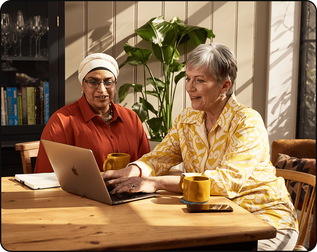 Two women sitting at a table using a laptop, each with a yellow mug, discussing something on the screen.